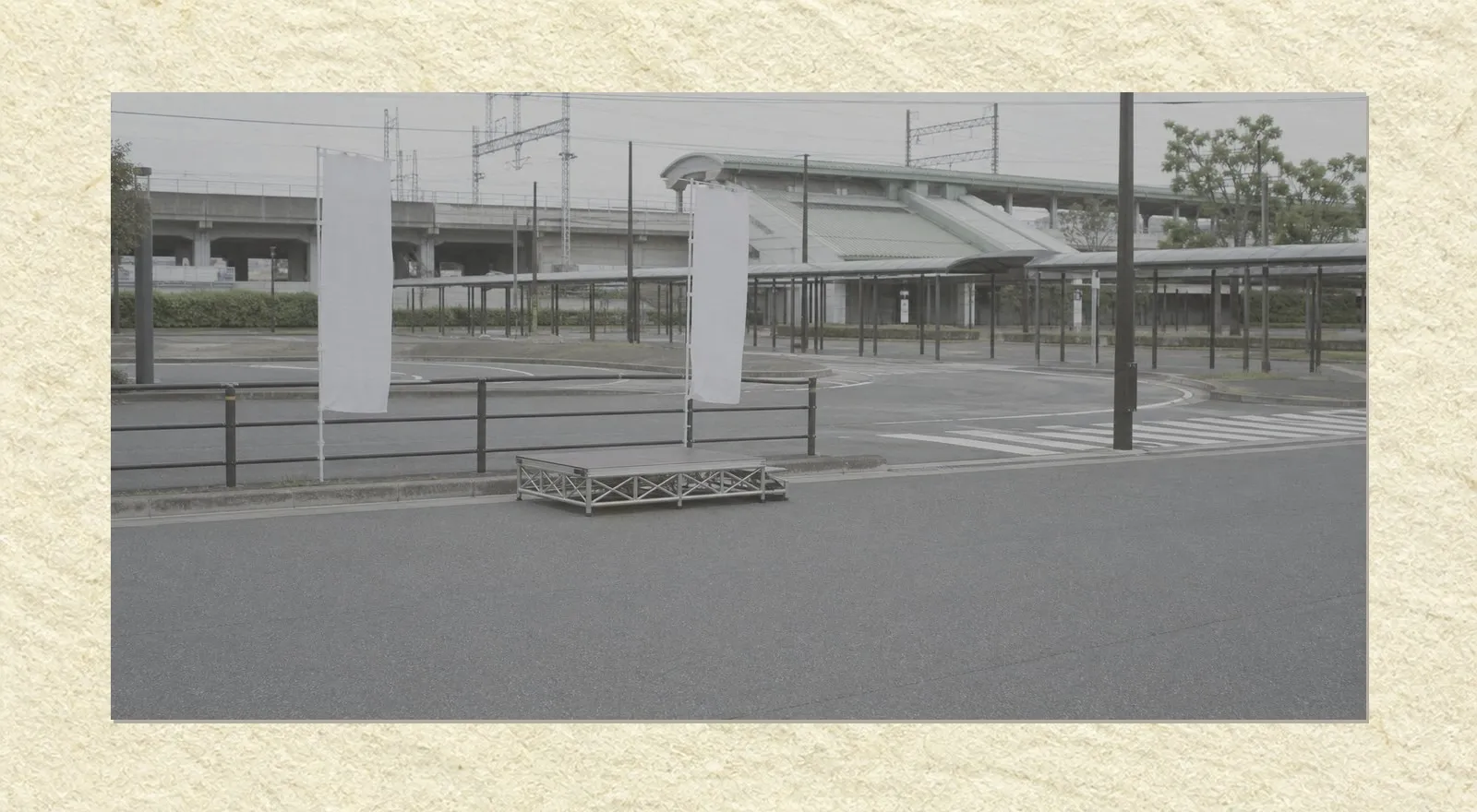 An empty stretch of road outside a suburban Japanese railway station under flat overcast light. A low portable speaking platform stands alone on the roadway beside a metal guardrail, with two blank banners on poles fixed to the guardrail. Beyond it an empty bus and taxi rotary and a faded pedestrian crossing. Nobody is present.