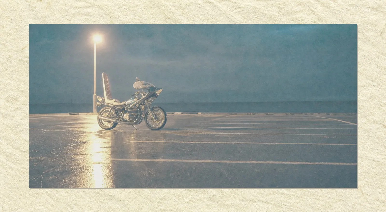 A deserted seaside parking area at night. One customised motorcycle with a tall backrest stands alone under a single street lamp, its chrome catching the light, the wet asphalt holding a long reflection. The rest of the lot is empty, marked only by faded white parking lines running into the dark. Nobody is present.
