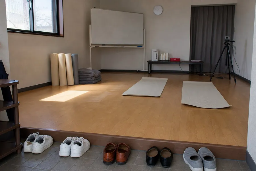 A photograph of a small rented room used for a yoga class, seen from the entrance step where five pairs of shoes are lined up: white sneakers, brown loafers, black flats and grey slip-ons. Inside, pale laminate flooring, two cream yoga mats still unrolled, rolled mats and folded blankets against the wall, a wiped whiteboard on a stand, a low table with a kettle, paper cups and a small red first-aid kit, a closed curtain across an alcove at the back, and a webcam on a tripod pointing at the empty floor. Late afternoon light falls from a window on the left. Nobody is present. AI-generated photo illustration; not a photograph of any real room, event or person.