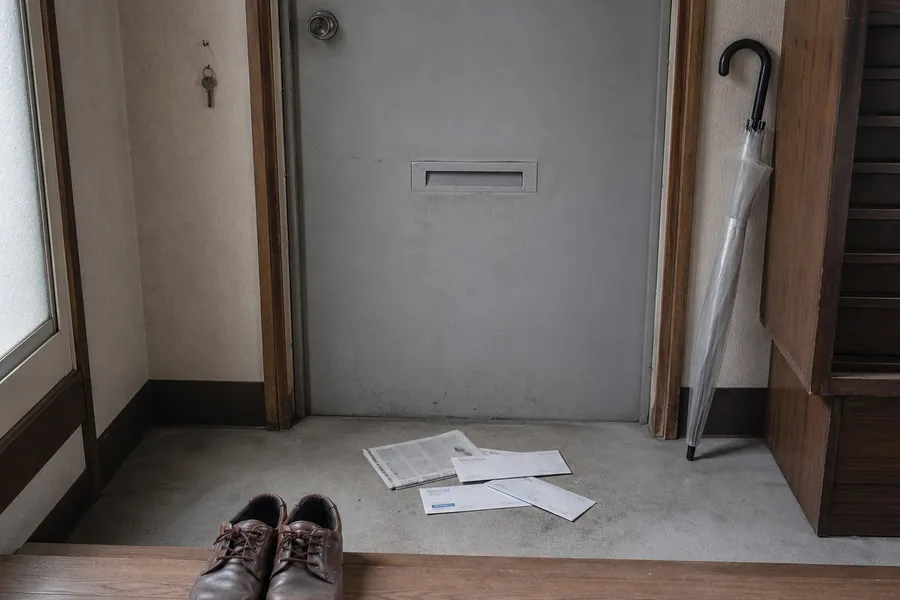 The inside of a small Japanese apartment entrance, photographed from within the room. A steel door with a letter slot stands closed. On the concrete floor of the sunken entrance below it lie a folded newspaper and several envelopes, left where they fell through the slot. A pair of men's worn leather shoes sits squared against the raised wooden step, toes pointing at the door. A clear plastic umbrella leans against the wall to the right. Weak daylight comes from a frosted window on the left. Nobody is present.