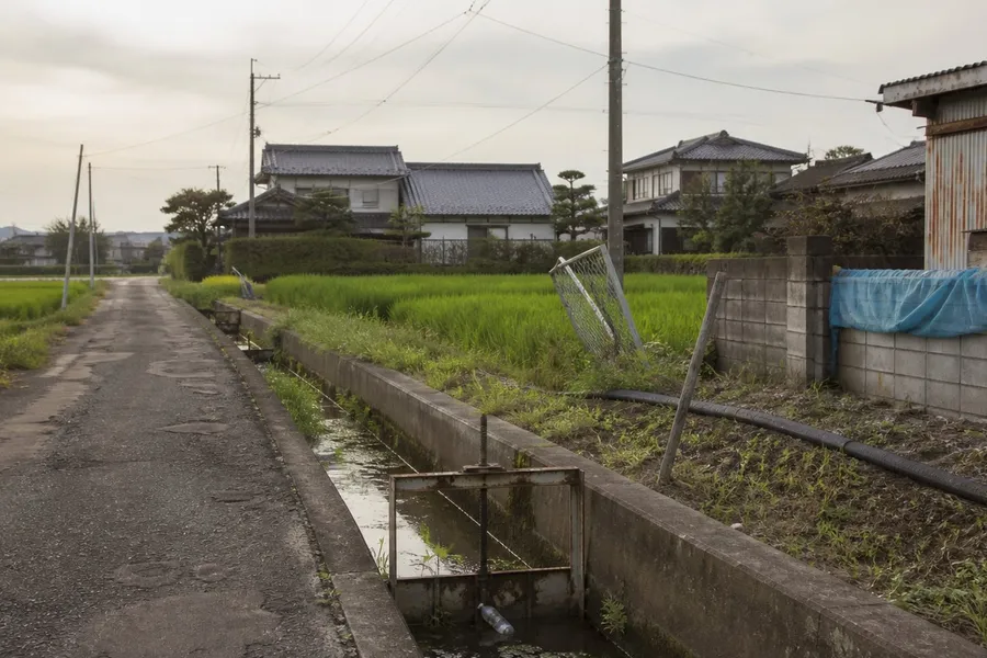 A photograph of the edge of a small Japanese country town in the late afternoon. A narrow asphalt farm road runs away from the camera on the left, its surface cracked and patched with tar, with no guardrail. Beside it a concrete-lined irrigation channel about a metre wide holds shallow standing water with patches of green algae and a small rusted metal sluice gate. Beyond the channel lie rice paddies at mid-growth, a bent galvanised fence panel, a black plastic irrigation pipe, concrete utility poles carrying bundled cables, a faded blue tarpaulin folded over a low concrete block wall, a corrugated iron shed, and small houses with grey tiled roofs. No people are present. AI-generated photo illustration; not a photograph of any real place, case or person.