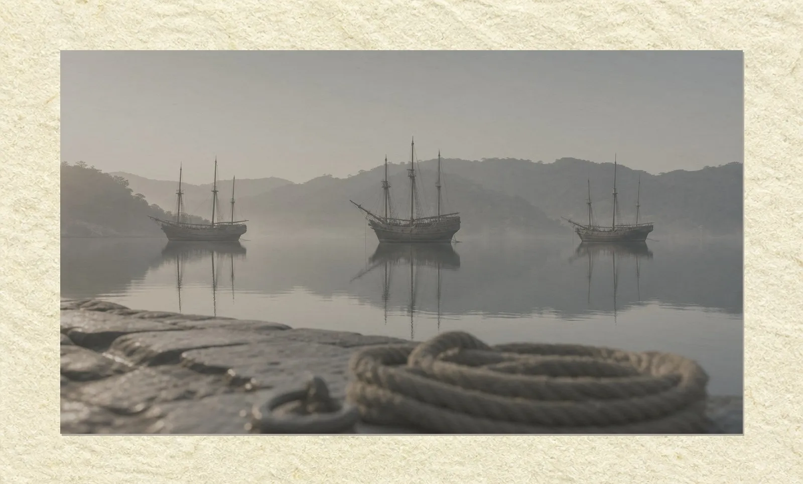 A wide, still harbour at first light, mounted on a sheet of pale paper with a narrow margin around it. Three tall wooden sailing ships lie at anchor in the middle distance with their sails furled, reflected in flat mirror-like water. Mist hangs over the surface and low wooded hills fade into haze behind. In the foreground, out of focus, the wet stone edge of a quay and a coiled rope. No people are present.