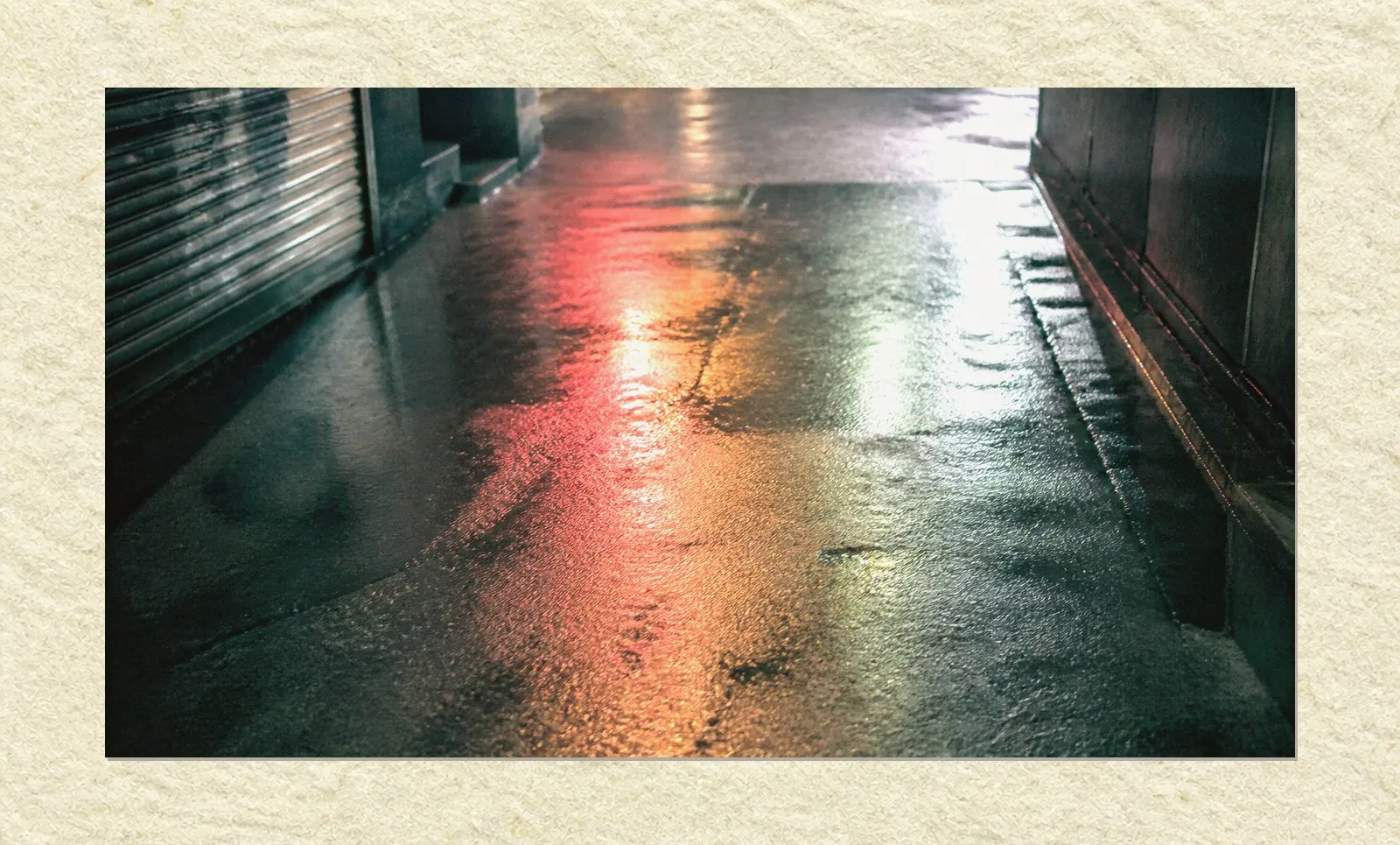 A narrow alley at night, seen looking down at the wet asphalt. Coloured light from signs somewhere above pools across the rain-slicked surface in red, amber and pale green. A closed metal shutter runs along one side. Nobody is present.
