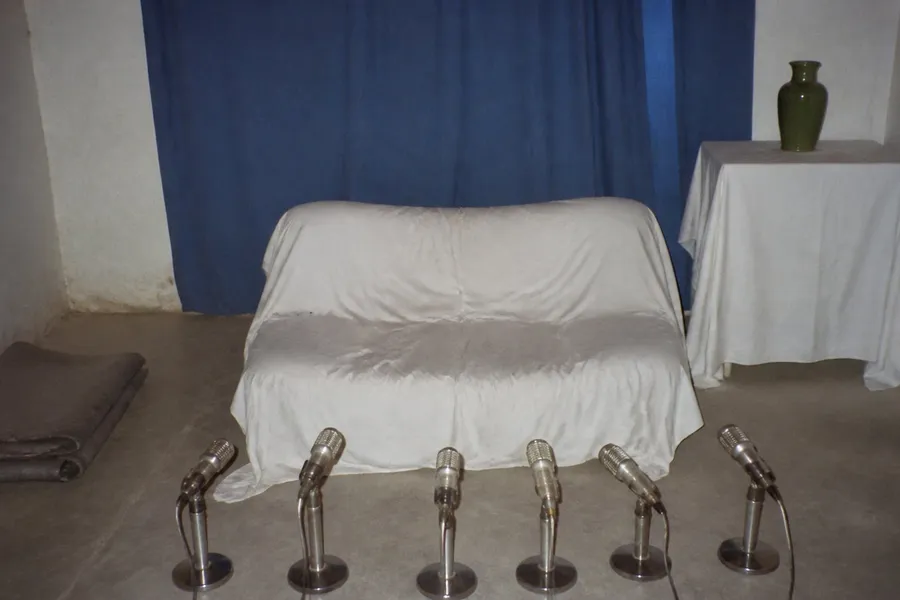 A photograph of a small plain room lit by direct camera flash, in the washed-out style of a 1990s amateur snapshot. At the centre a low couch draped in white cloth stands empty, the cloth still creased and dented where somebody had been sitting. Facing the empty seat, a row of six old silver microphones on short stands is lined up along the bare floor. A plain blue cloth curtain hangs slightly crooked on the wall behind, a low white-clothed table at one side holds a green ceramic vase, and a folded grey blanket lies on the floor. Nobody is present. AI-generated photo illustration; not a photograph of any real room, event or person.