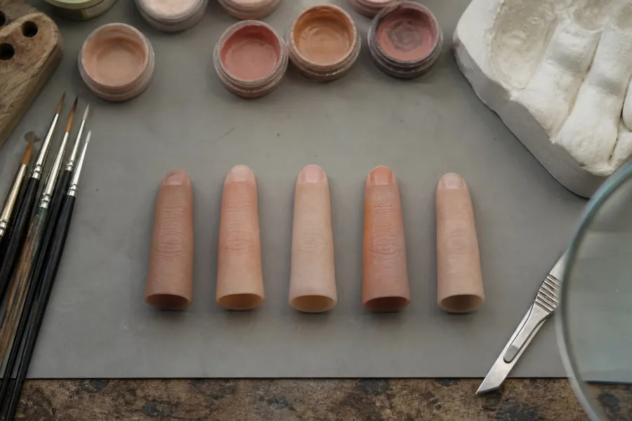A photograph looking down on the workbench of a small prosthetics workshop. Five silicone prosthetic fingertips in slightly different skin tones lie in a row on a grey work mat, each hollow and open at its base. Around them are fine sable brushes, open pots of skin pigment, a scalpel, a plaster cast of a hand and the edge of a magnifying lamp. Everything is worn and orderly. No people and no hands appear. AI-generated photo illustration; not a photograph of any real workshop.