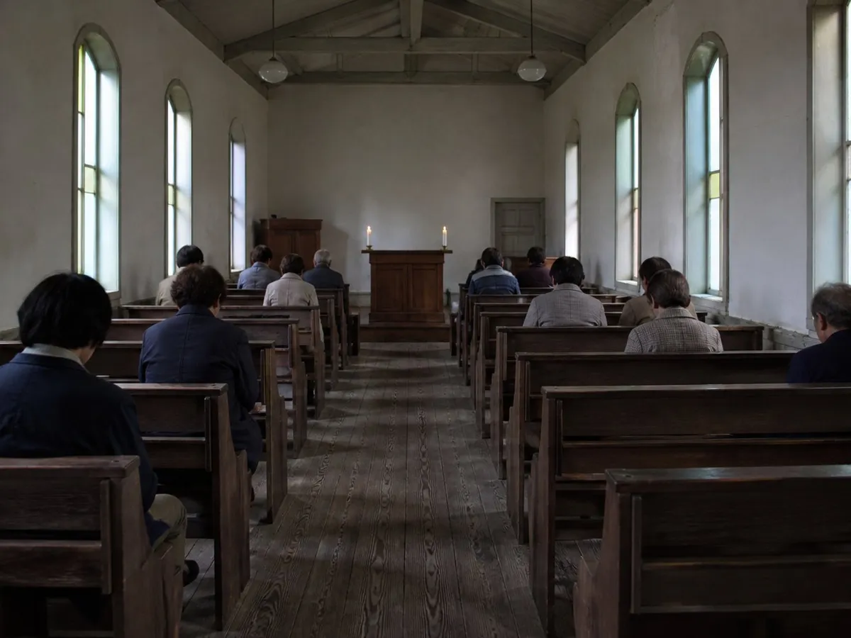 A photograph of the interior of a very small old Christian church in western Japan, taken from the back of the nave during a service. Ten or twelve people, most of them elderly, sit far apart on plain dark wooden pews, all seen from behind with heads slightly bowed; the great majority of the pews are empty. Whitewashed plaster walls, a worn plank floor, exposed ceiling beams, tall narrow windows of pale coloured glass, and a simple wooden table with two candles at the far end. Modest and poor but carefully kept. No faces are visible. AI-generated photo illustration; not a photograph of any real church, service or person.