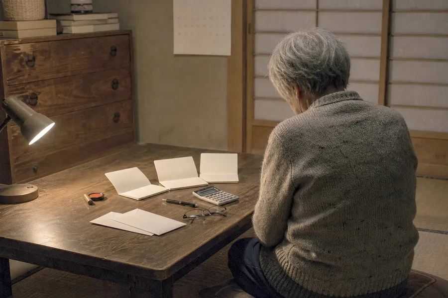 A photograph of a dim tatami room in an older Japanese house late at night, lit only by a single desk lamp. On a low dark wooden table lie three bank passbooks opened flat, a small cylindrical personal seal beside a red ink pad, two plain white envelopes, a pocket calculator, a pen and a folded pair of reading glasses. An elderly woman in a plain knitted cardigan kneels at the table with her back to the camera, her head slightly bowed and her hands resting in her lap; no face is visible. A paper sliding screen, a low chest of drawers and a blank wall calendar stand behind her, and the corners of the room fall away into shadow. AI-generated photo illustration; not a photograph of any real person or household.